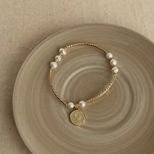 Pulsera de cuentas doradas y perlas con una medalla grabada colgando.