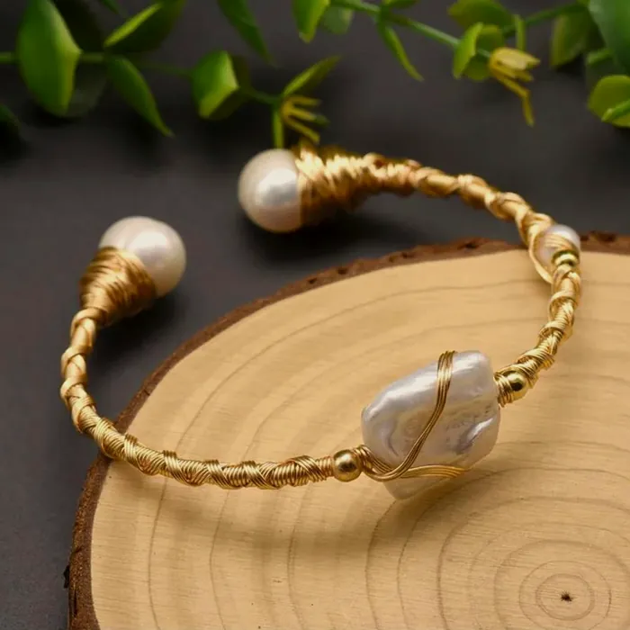 Brazalete rígido de cuentas doradas abierto, con una perla barroca en cada extremo, sobre madera.