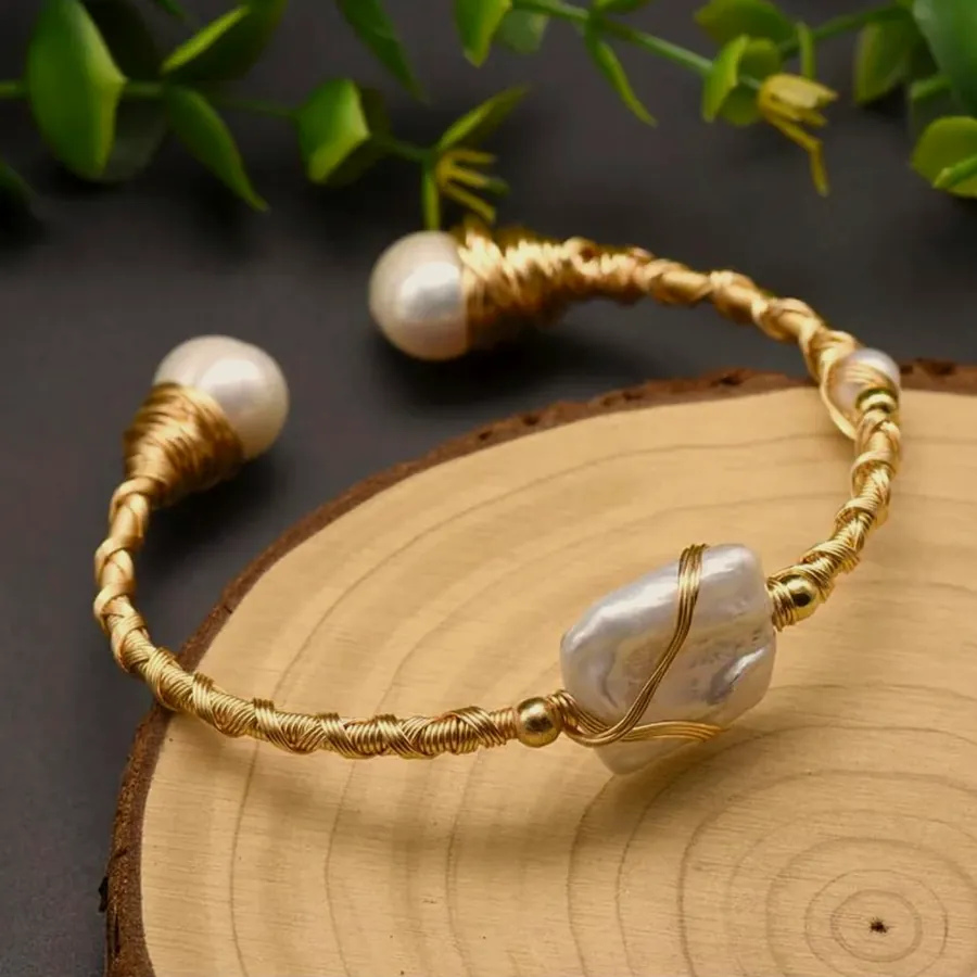 Open rigid cuff of gold-tone beads with a baroque pearl at each end, on wood.