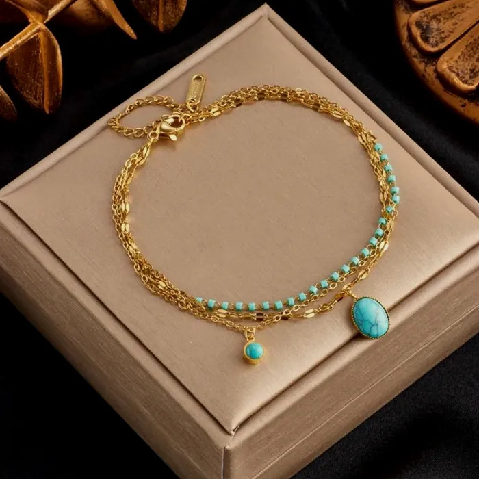 Pulsera de doble vuelta con cadena dorada y cuentas de turquesa, con una turquesa colgando, en su caja.